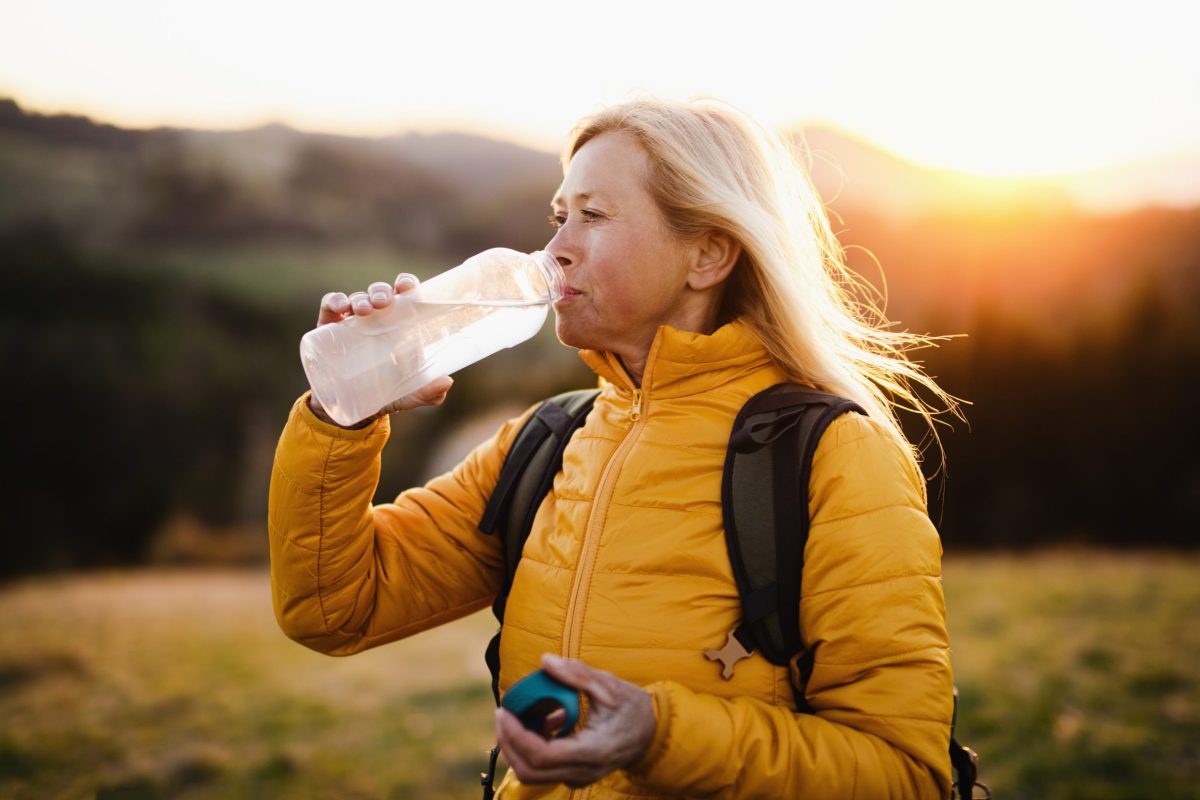 Attractive senior woman hiker walking outdoors in nature at sunset, drinking water.